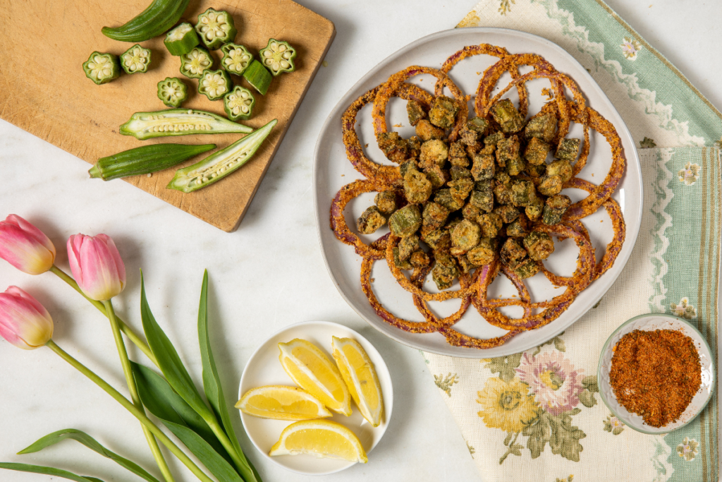 Plate of crispy fried okra and onion rings on white plater on patterned cloth, surrounded by sliced okra, seasoning, lemon wedges, and pink tulips on a white surface.