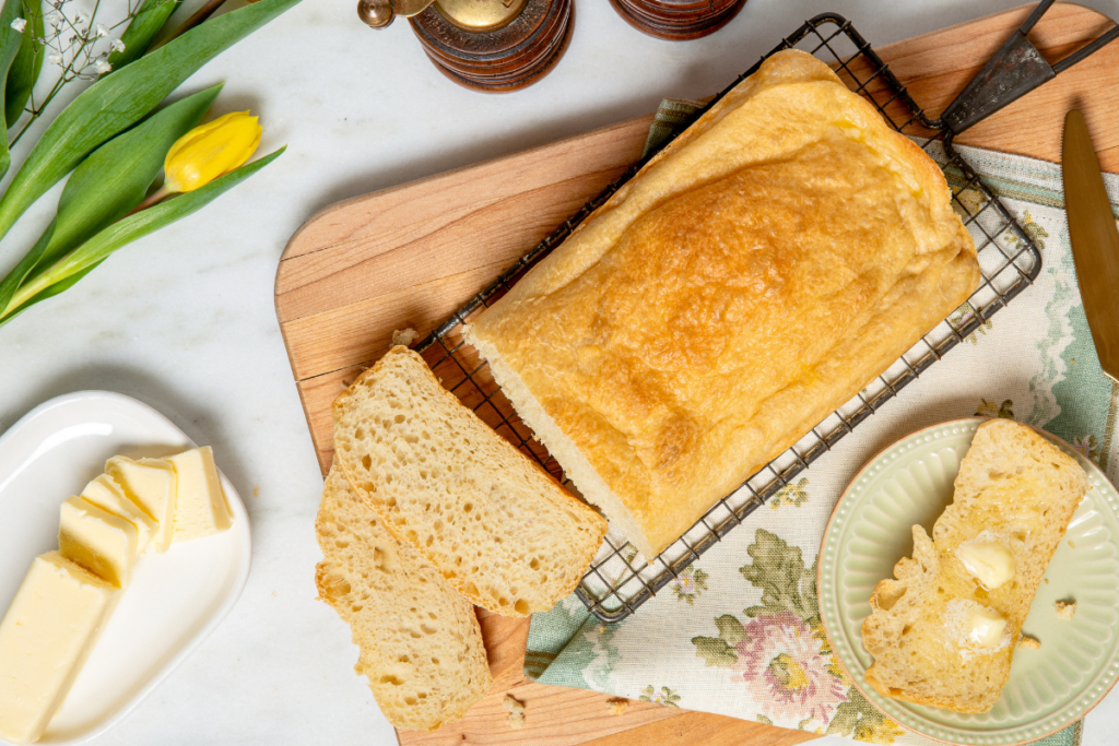 Golden homemade bread loaf on a cooling rack with buttered slices, butter dish, and a yellow tulip stem on a floral cloth.