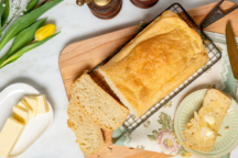 Golden homemade bread loaf on a cooling rack with buttered slices, butter dish, and a yellow tulip stem on a floral cloth.