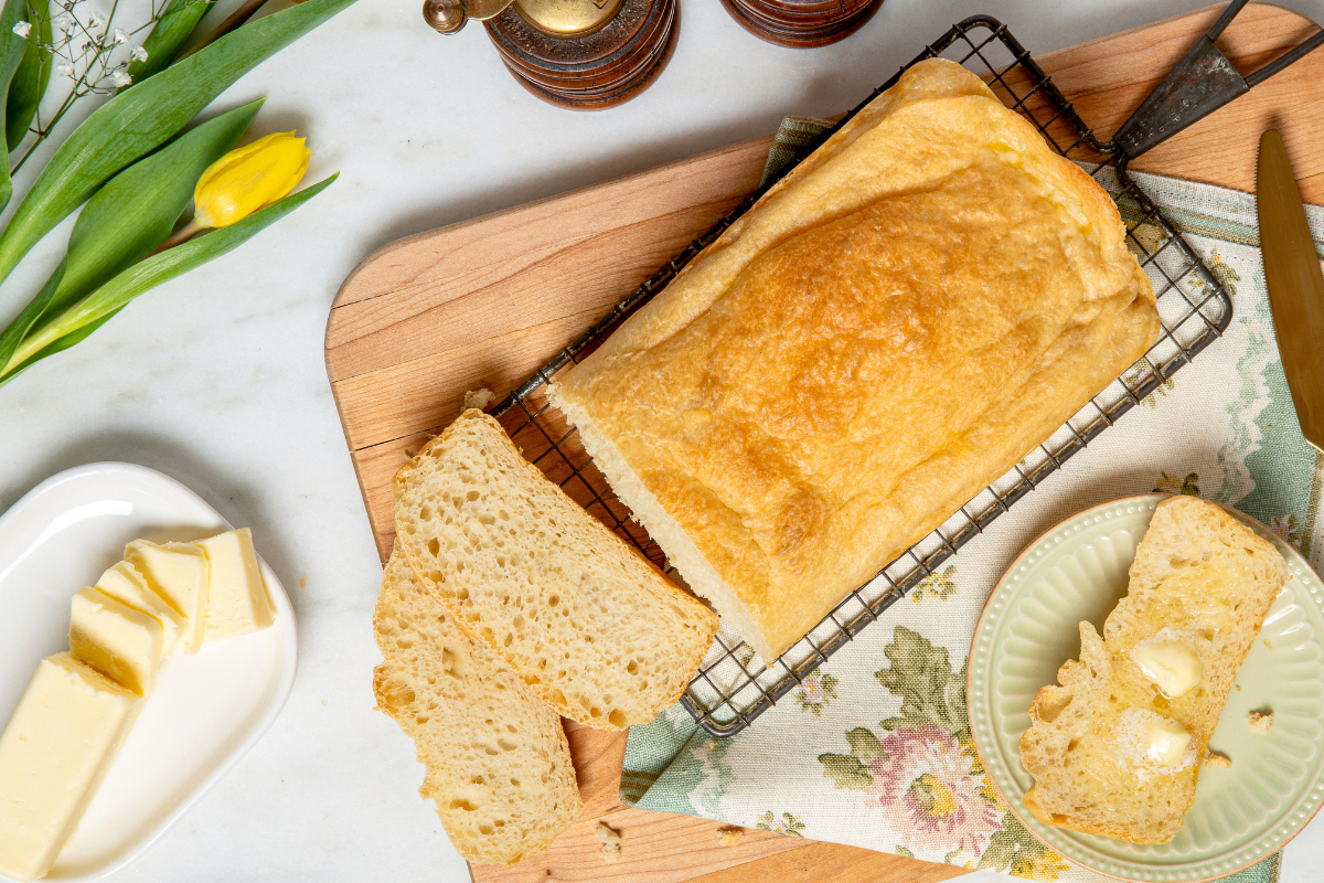 Golden homemade bread loaf on a cooling rack with buttered slices, butter dish, and a yellow tulip stem on a floral cloth.