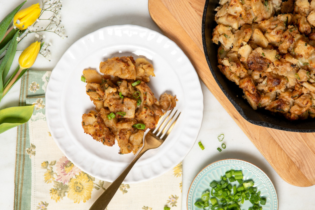 Crispy smothered potato served in a cast iron skillet and white plate with green onion garnish and yellow tulips on the side.