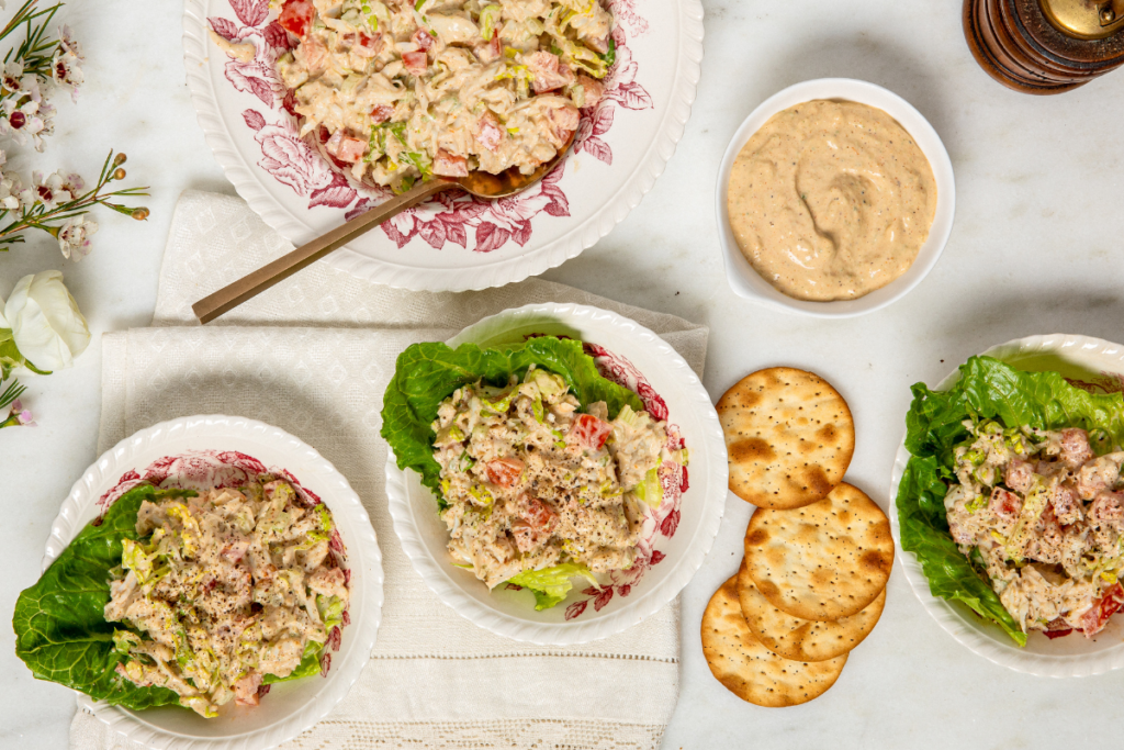 Three creamy crab salad bowls served on bed of lettuce and a larger serving bowl, accompanied by crackers and a side of dressing