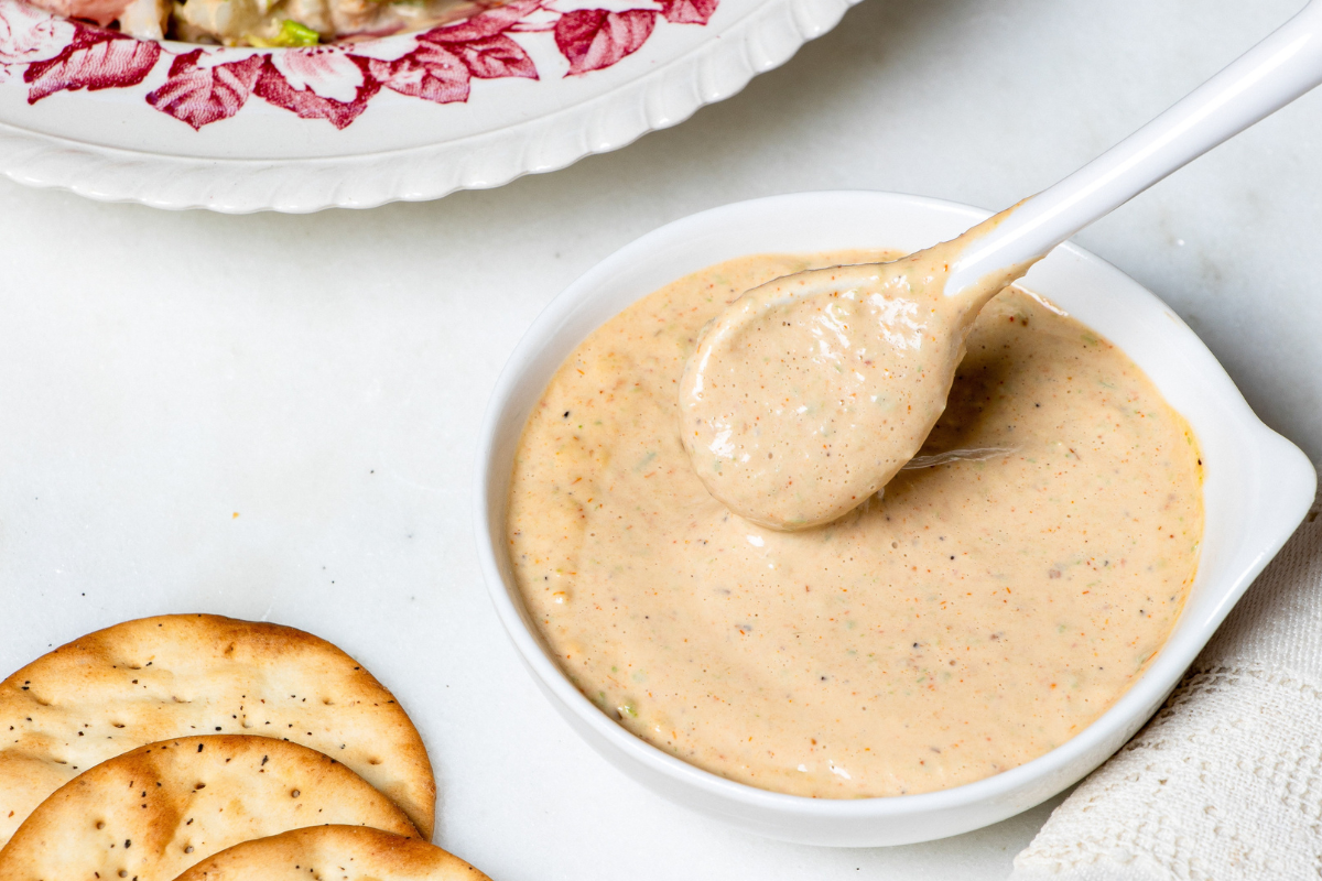Close-up of a creamy sauce being lifted with a spoon from a small white bowl, with crackers visible alongside