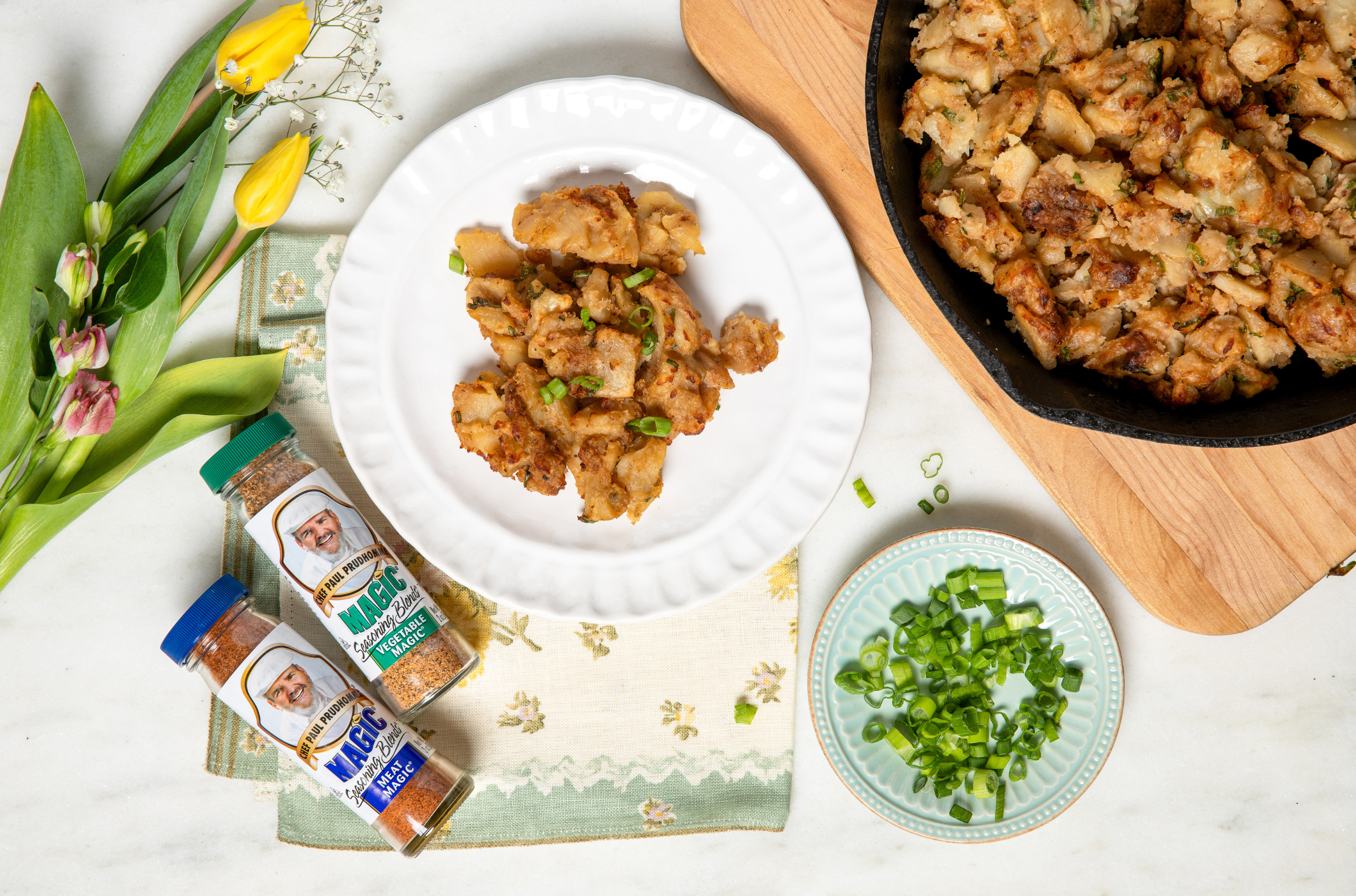 Plate of golden fried potatoes garnished with green onions beside seasoning bottles and fresh flowers, creating a warm and appetizing scene.