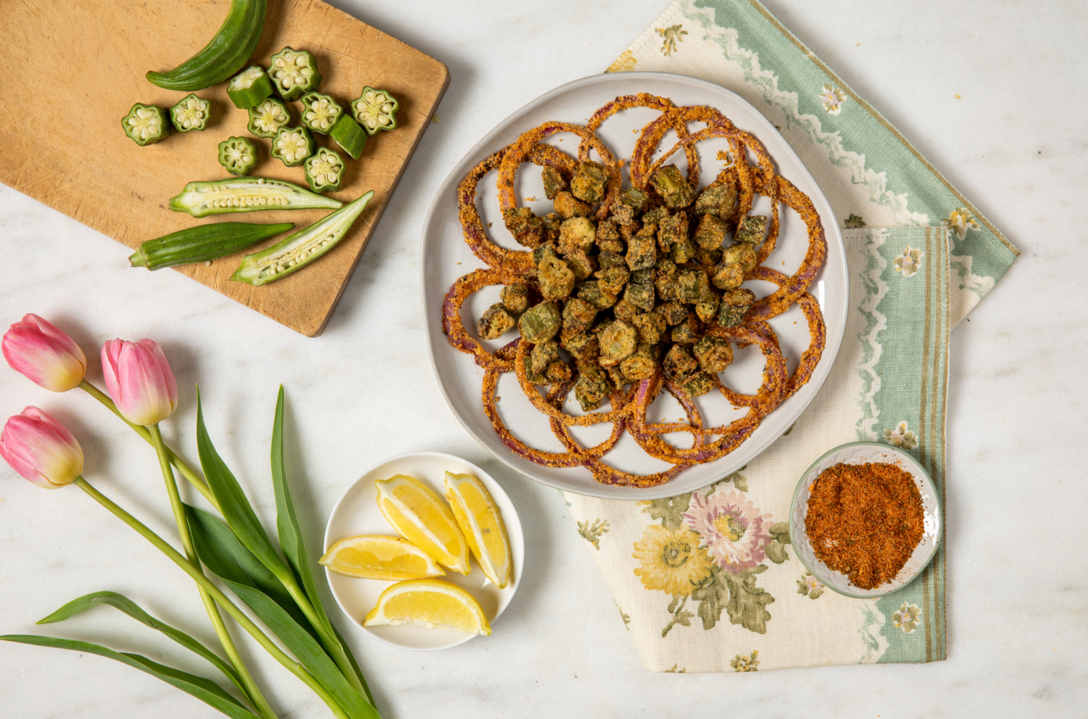 Plate of crispy fried okra and onion rings on white plater on patterned cloth, surrounded by sliced okra, seasoning, lemon wedges, and pink tulips on a white surface