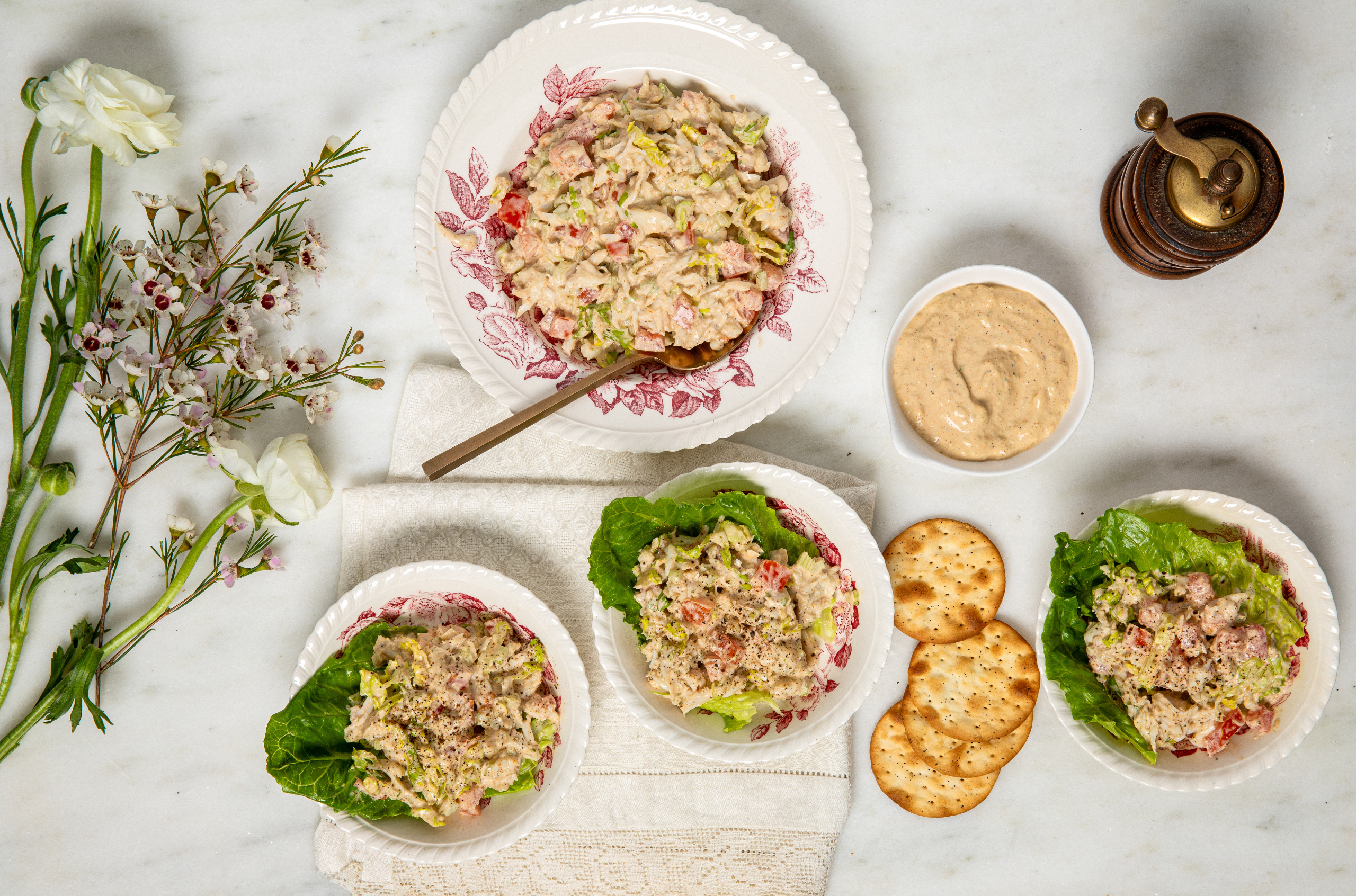 Aerial view of a elegant table setting with chicken salad on leafy greens, surrounded by crackers, flowers, a dip bowl, and a pepper mill, on a marble background.
