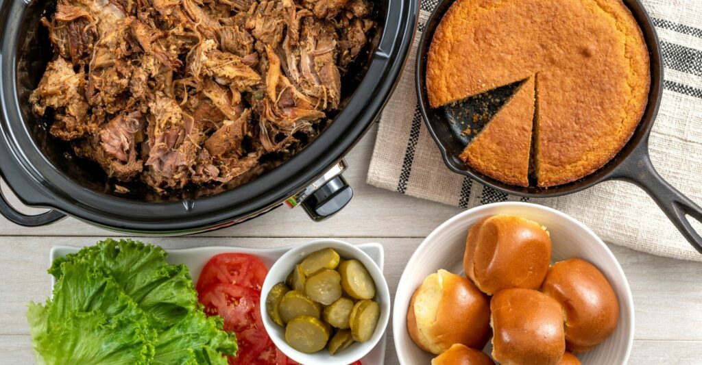 a slow cooker full of slow cooker cooked cochon de lait, next to a cast iron skillet of corn bread with a piece taken out, a bowl of rolls and a tray of various fixings including lettuce, pickles and tomatoes