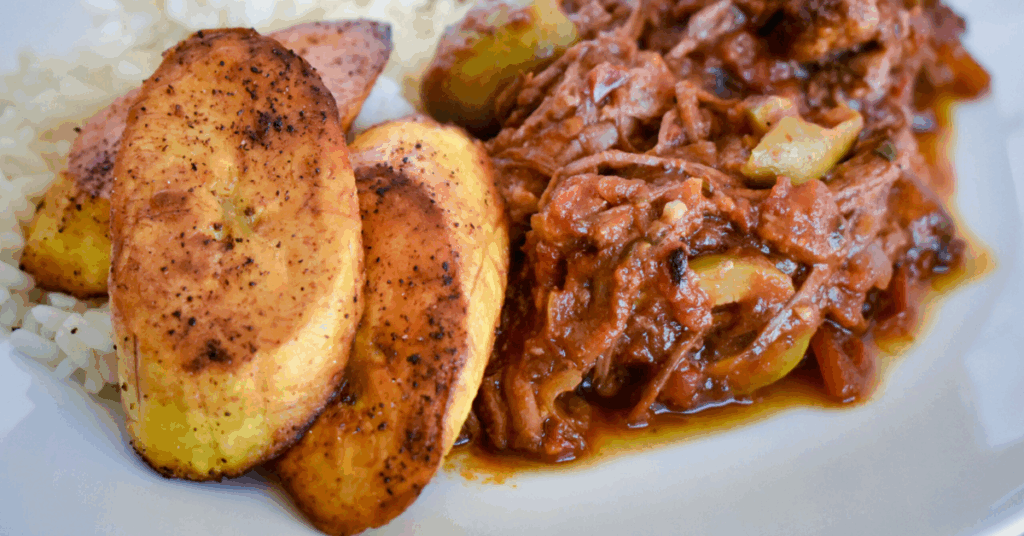 A dish containing cooked meat paired with fluffy rice and plantains on a white plate.