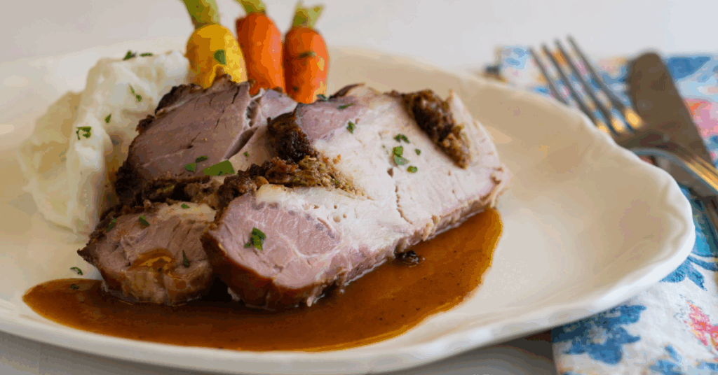 Roasted pork slices with gravy, mashed potatoes, and colorful carrots on a white plate. A fork and knife rest on a floral napkin to the side.