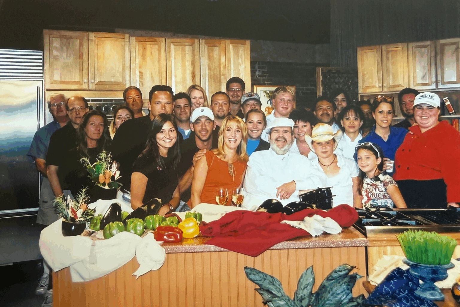 This is a group photo taken in a kitchen setting, featuring a diverse crowd of people smiling and gathered closely together. In the front, there are several individuals, including a man in a white chef's coat and hat, and a woman with blonde hair in an orange dress, both holding glasses of wine. The table in front of them displays colorful vegetables, flowers, and a red cloth, suggesting a celebration or a culinary event. The background shows wooden kitchen cabinets and modern appliances, creating a warm and lively atmosphere.