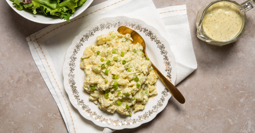 A plate of fresh potato salad drizzled with dressing, with a bowl of vibrant greens on the side and an extra bowl of dressing nearby.