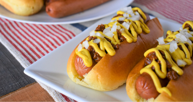 Two hot dogs on a white plate topped with chili and dressed with onions and mustard. 