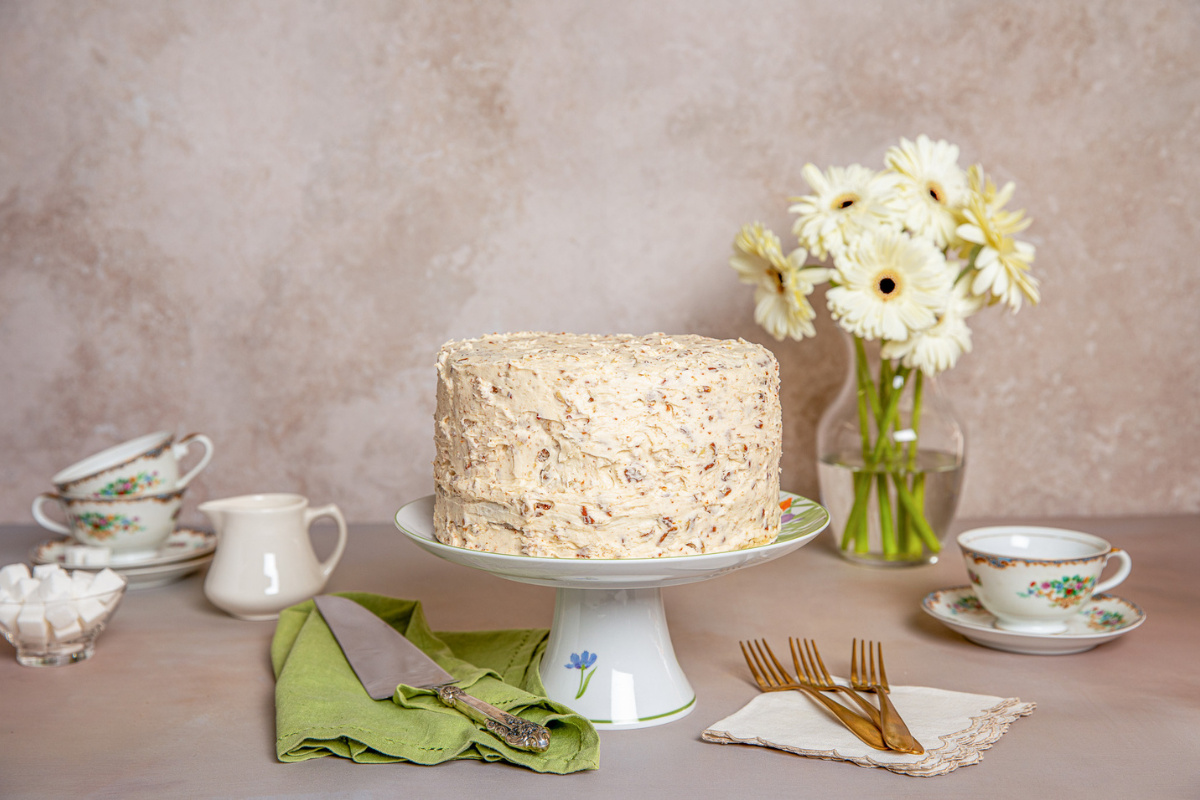 A textured cake on a floral stand, surrounded by white daisies, elegant teacups, a creamer, sugar cubes, and gold forks, creating a vintage tea party vibe.