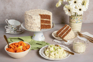 A round Spiced Pecan Cake with Pecan Frosting cake on a plate with a slice removed, revealing its layered interior and frosting. Surrounded by a bowl of sweet potatoes and a plate of potato salad.