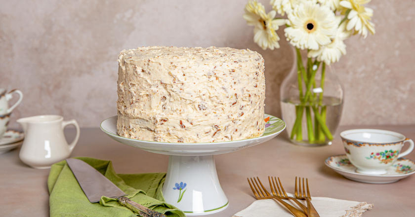 A textured cake on a floral stand, surrounded by white daisies, elegant teacups, a creamer, sugar cubes, and gold forks, creating a vintage tea party vibe.