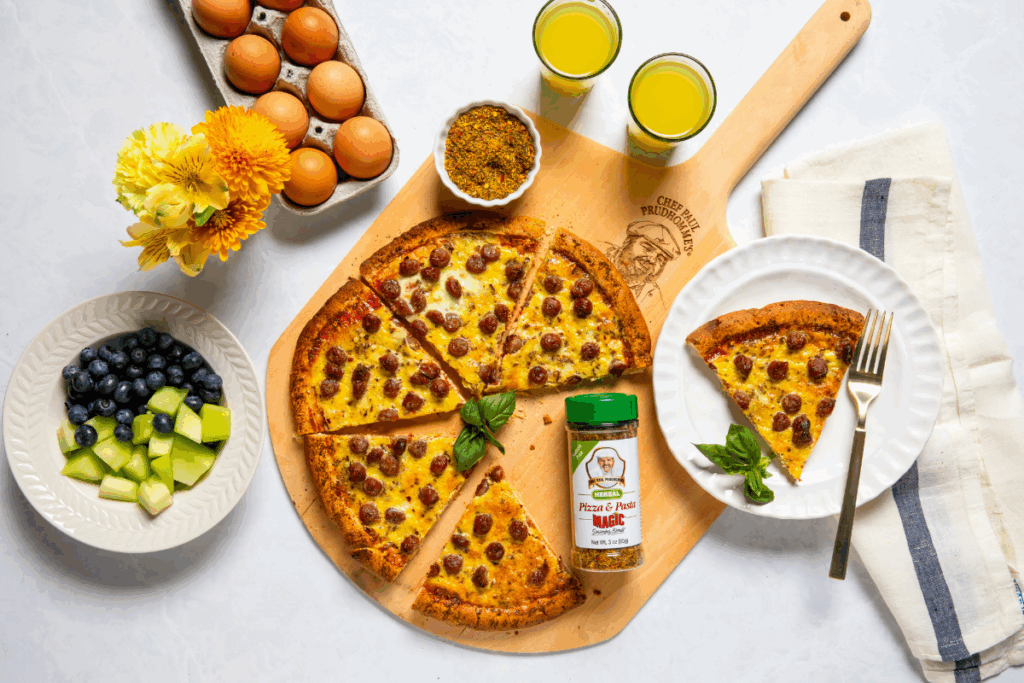 Pizza on a wooden board, sliced with basil garnish. Surrounding items: carton of eggs, bowl of honeydew and blueberries, flowers, spice jar, and orange juice.