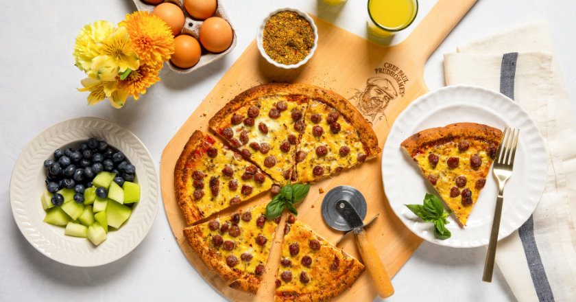 A sausage pizza on a wooden board, partially sliced, with a pizza cutter. Nearby are blueberries, melon, eggs, flowers, and a glass of juice, creating a fresh, inviting breakfast setup.
