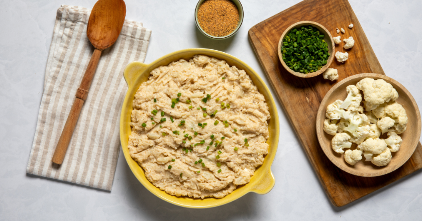 Mashed cauliflower in a yellow dish, garnished with chives. Surrounding are bowls of chives, cauliflower florets, seasoning, a wooden spoon, and a striped napkin.