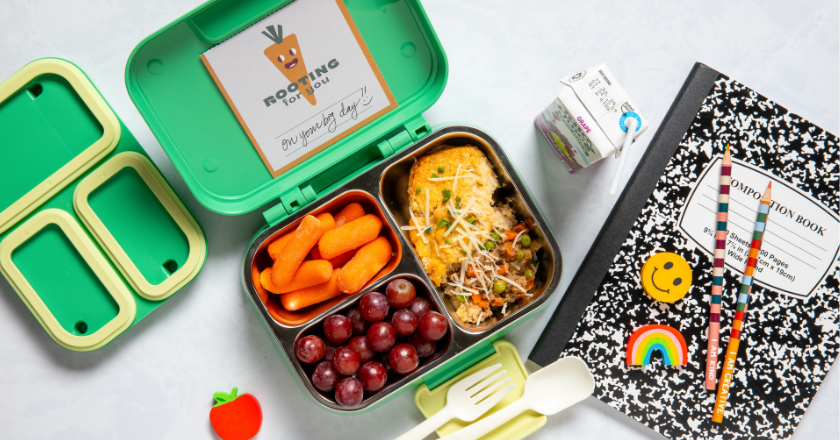Green lunchbox with a note, filled with carrots, grapes, and an omelette. Nearby are a milk carton, utensils, a notebook, and colorful pencils.