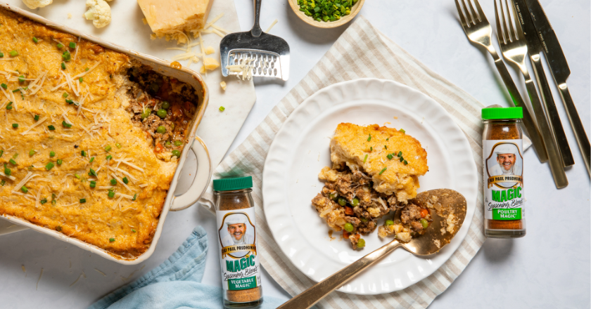 A casserole dish with a cheesy golden topping sits near a plate with a serving. Seasoning bottles, herbs, and utensils are displayed around, evoking a cozy, home-cooked meal.