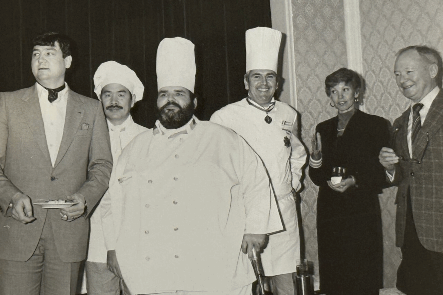 A group of people stands together indoors. Three chefs in white uniforms and tall hats are flanked by a man in a suit and a woman holding a drink, smiling.