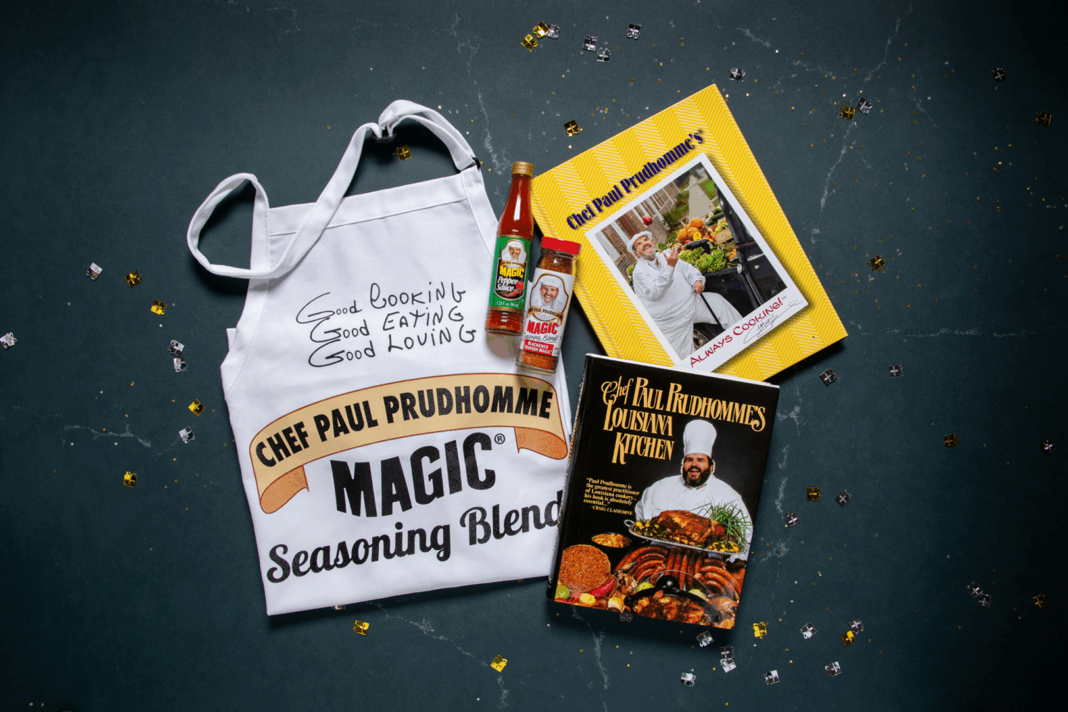 Apron with "Good Cooking, Good Eating, Good Loving" and "Chef Paul Prudhomme Magic Seasoning Blend," hot sauce, seasoning bottle, and two cookbooks on dark background. Festive and culinary-themed layout.