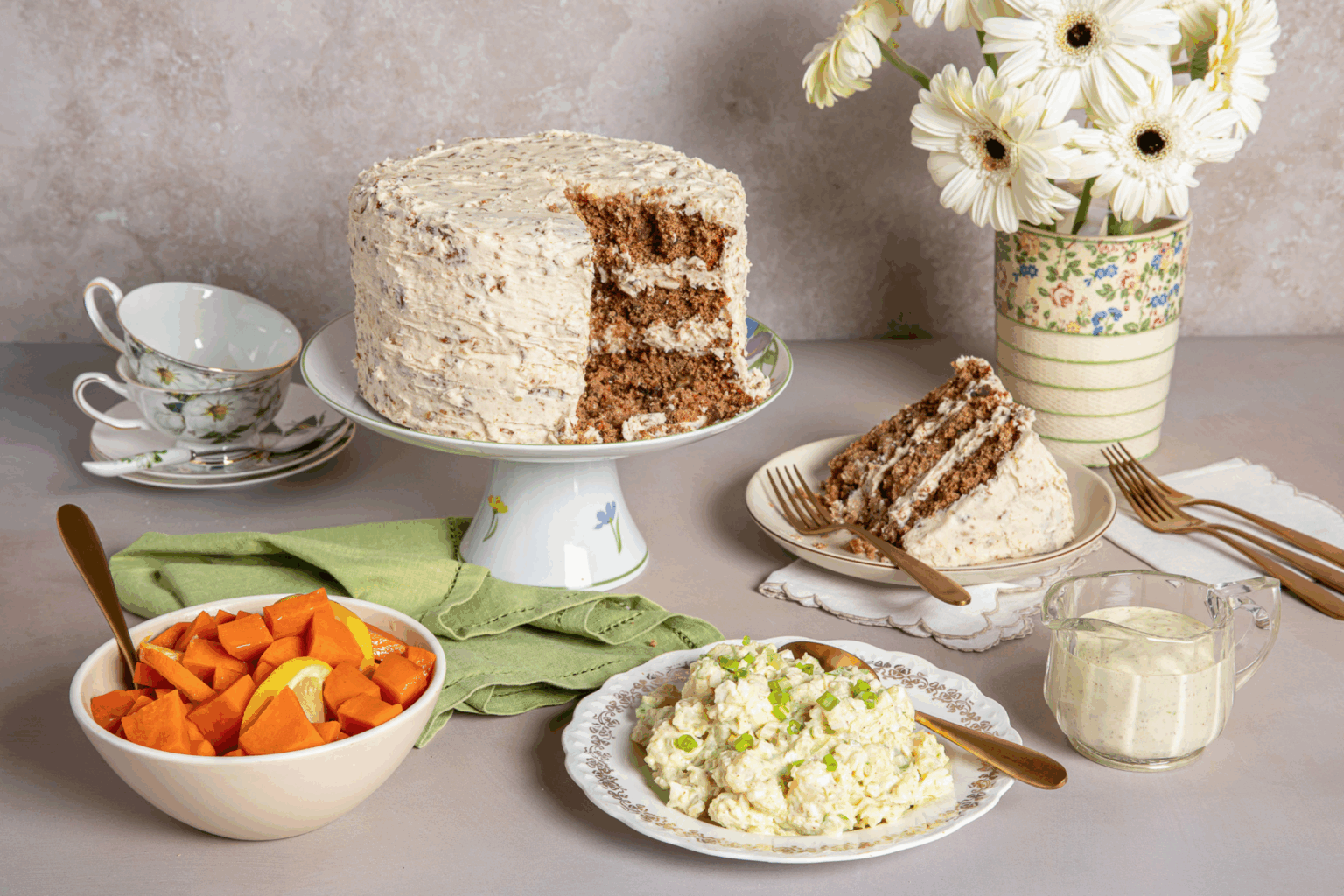 A frosted cake on a stand with a slice on a plate beside it, surrounded by a bowl of sweet potatoes, potato salad, daisy-decorated mugs, and white flowers.