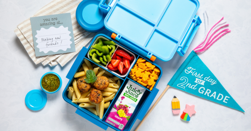 Blue lunchbox with pasta and meatballs, sliced strawberries, green bell peppers, fish-shaped crackers, and grape juice. Nearby: card with motivational note, pesto dip, and turquoise "First day of 2nd Grade" pennant. Bright, cheerful tone.