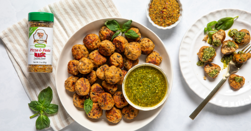 Plate of browned meatballs garnished with basil, served with green pesto sauce. A spice jar is nearby on a striped cloth. Mood is appetizing and fresh.