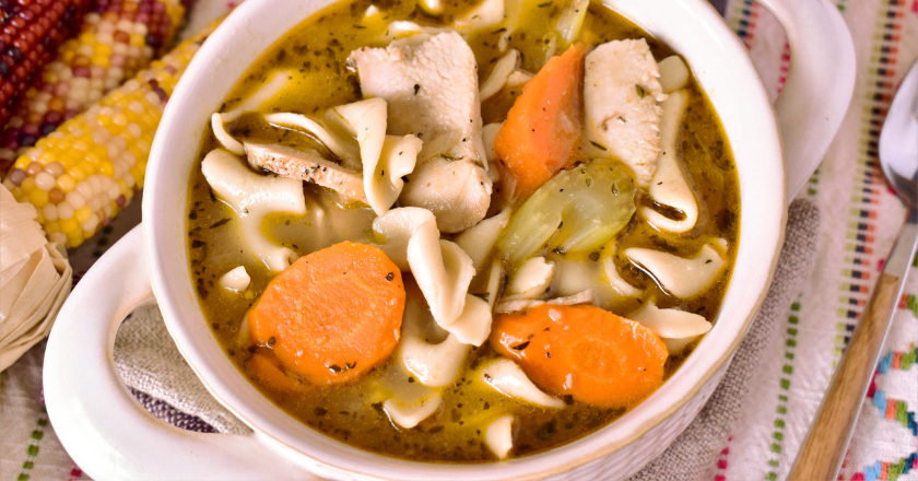 A bowl of chicken noodle soup with carrots and celery, garnished with herbs. It conveys warmth and comfort. Colorful corn cobs in the background.