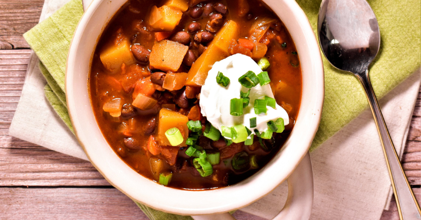 A bowl of hearty chili with black beans, diced vegetables, and a dollop of sour cream topped with chopped green onions. A spoon and lime wedges are nearby.