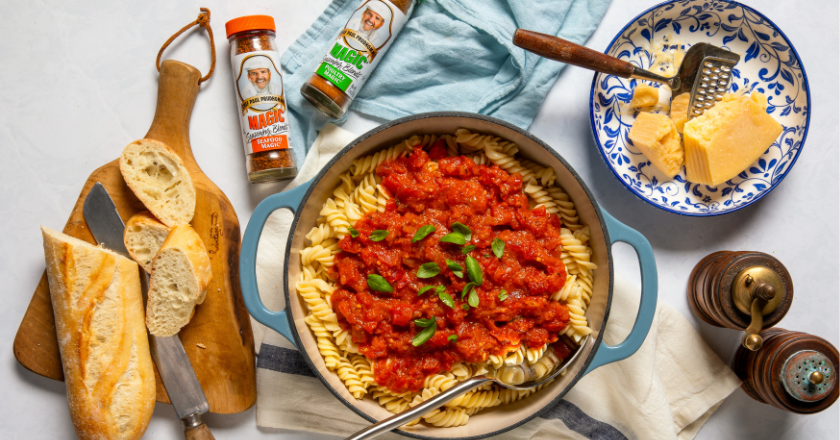 A bowl of fusilli pasta topped with red tomato sauce and basil. There's sliced baguette on a board, grated cheese, seasonings, and a blue towel nearby.