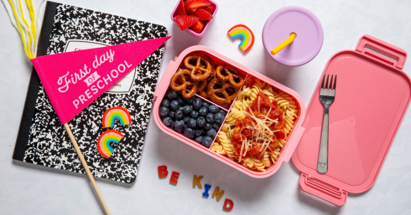 Pink lunchbox with pretzels, blueberries, pasta, near strawberries in a cup. A notebook reads “First day of preschool.” Colorful magnets spell “BE KIND.”