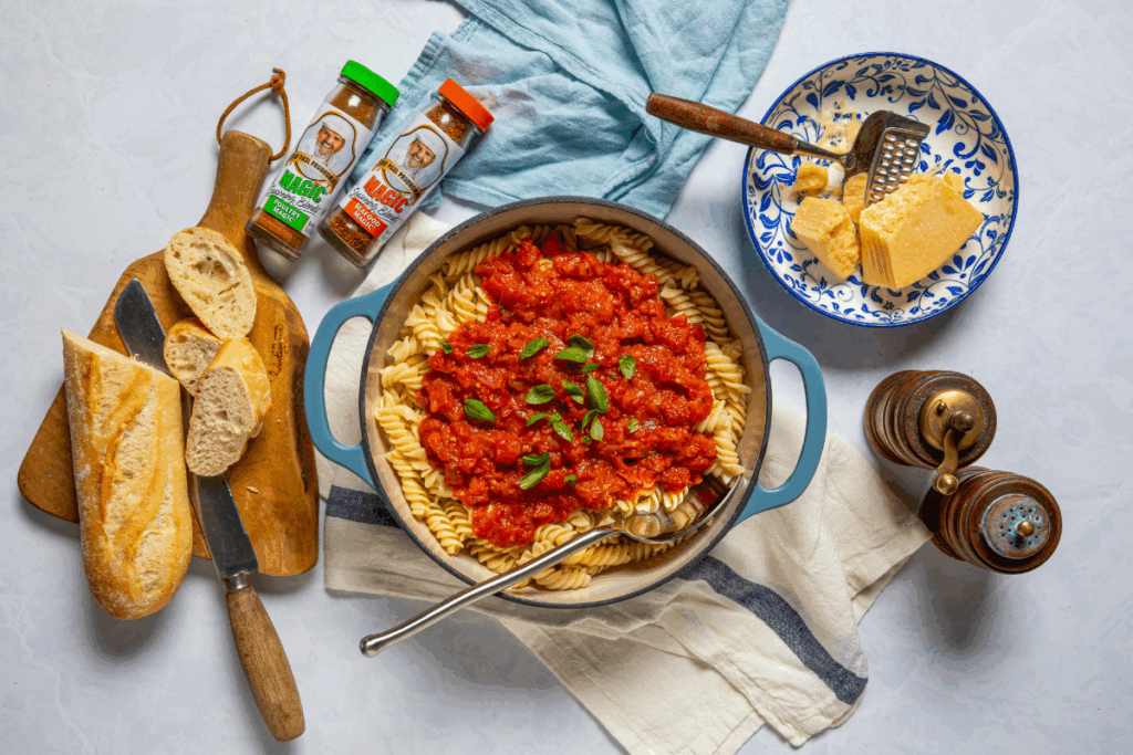 A pot of fusilli pasta topped with tomato sauce and basil sits on a cloth. Nearby are sliced bread, a cheese bowl with a grater, spice jars, and pepper mills.
