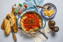 A pot of fusilli pasta topped with tomato sauce and basil sits on a cloth. Nearby are sliced bread, a cheese bowl with a grater, spice jars, and pepper mills.