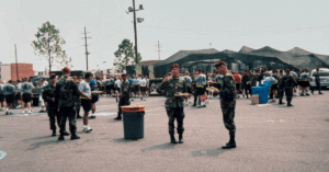 Soldiers in camouflage uniforms and berets gather outdoors on a warm day, holding plates of food. A large group of people is seen in the background, standing near a camouflaged tent. The atmosphere appears relaxed and social. Trees, utility poles, and scattered vehicles provide a casual setting.