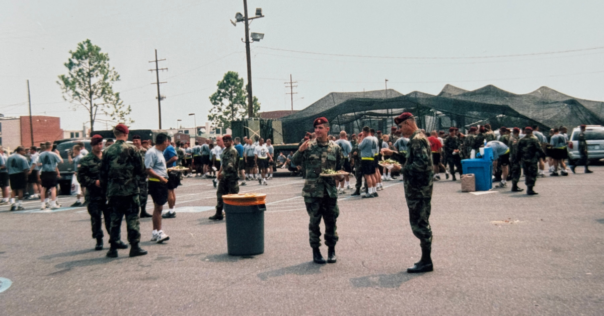 Soldiers in camouflage uniforms and berets gather outdoors on a warm day, holding plates of food. A large group of people is seen in the background, standing near a camouflaged tent. The atmosphere appears relaxed and social. Trees, utility poles, and scattered vehicles provide a casual setting.
