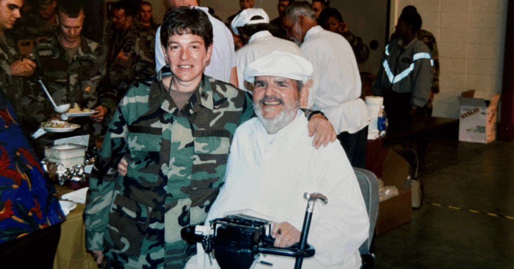 A person in a military uniform stands beside and embraces Chef Paul. The setting is a bustling dining hall with people in the background enjoying a meal, creating a warm, communal atmosphere.