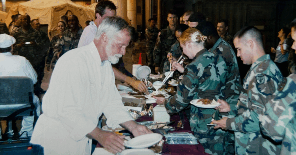 A bearded man in a white apron serves food to smiling military personnel in camouflage uniforms, creating a warm and convivial atmosphere.