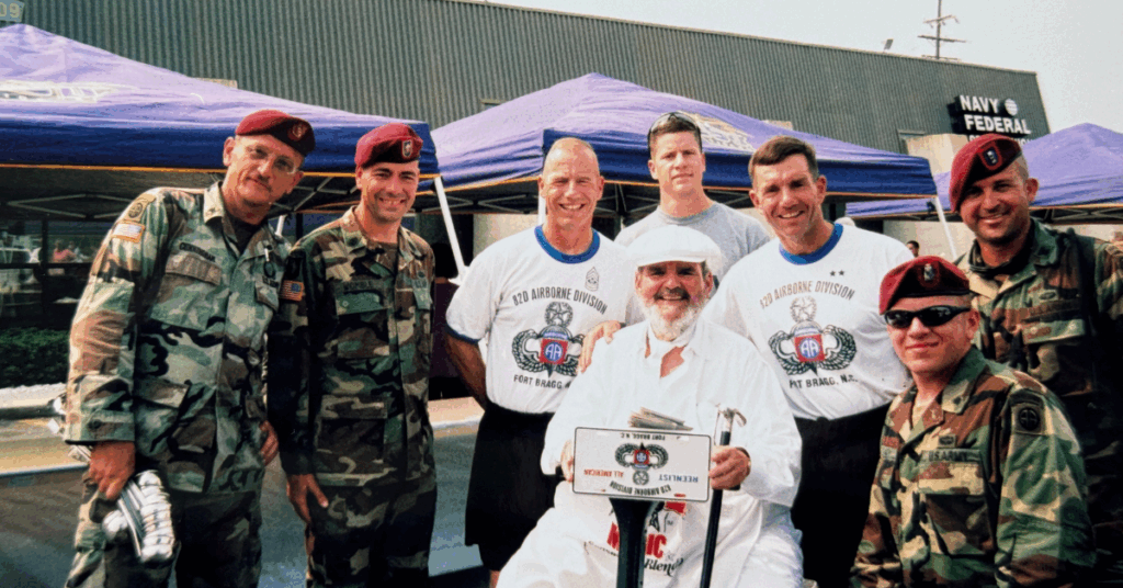 A group of military personnel in camouflage and red berets pose with Chef Paul. They stand in front of blue tents, smiling warmly.