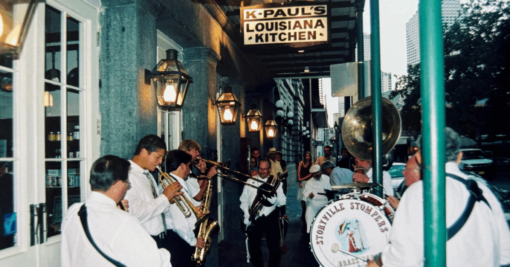 Street jazz band performing outside K-Paul's Louisiana Kitchen. Musicians play brass instruments energetically, creating a lively and festive atmosphere.