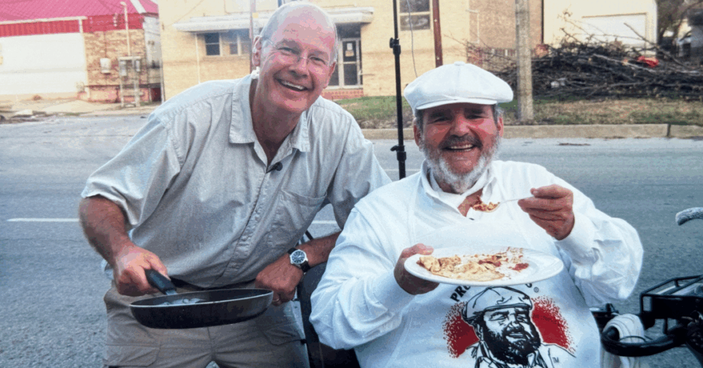 Two people smiling outdoors, one holding a pan and the other in a white apron and cap eating from a plate. The background shows urban buildings.