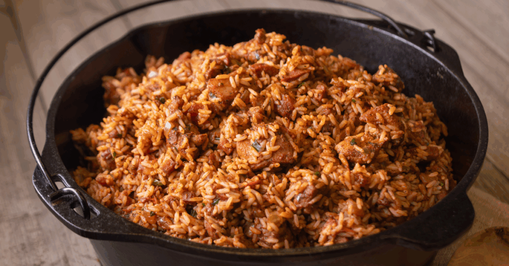 A black pot on a wooden table filled with spicy reddish-brown jambalaya, featuring rice, sausage, and herbs, evoking a warm, rustic meal.