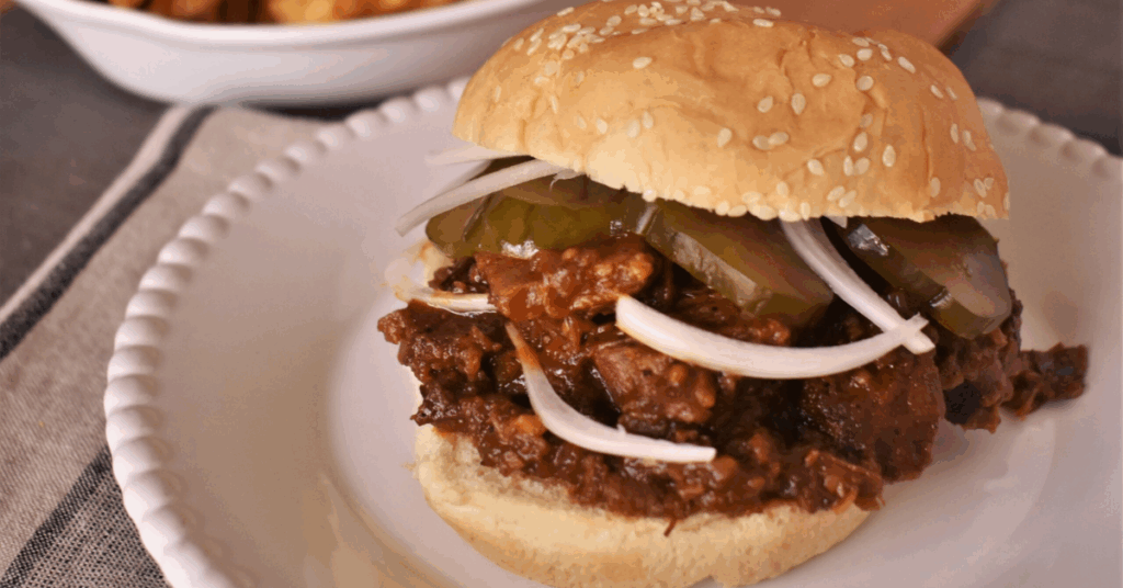 Game Day - A sloppy joe sandwich with onion slices and pickles on a sesame seed bun, served on a white plate. Waffle fries are in a bowl in the background.