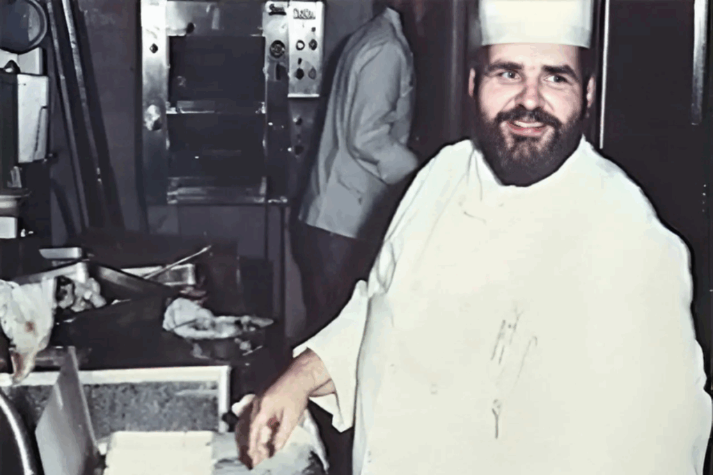 A bearded chef wearing a white uniform and hat smiles while working in a busy kitchen. Stainless steel appliances and another staff member are visible.