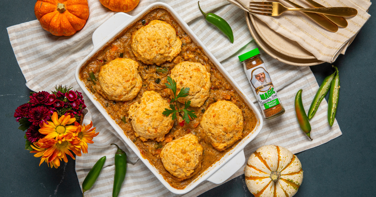 A casserole dish with golden biscuits on a stew-like base is on a striped cloth. Surrounded by a spice jar, green chilies, mini pumpkins, flowers, and plates. Cozy and autumnal.