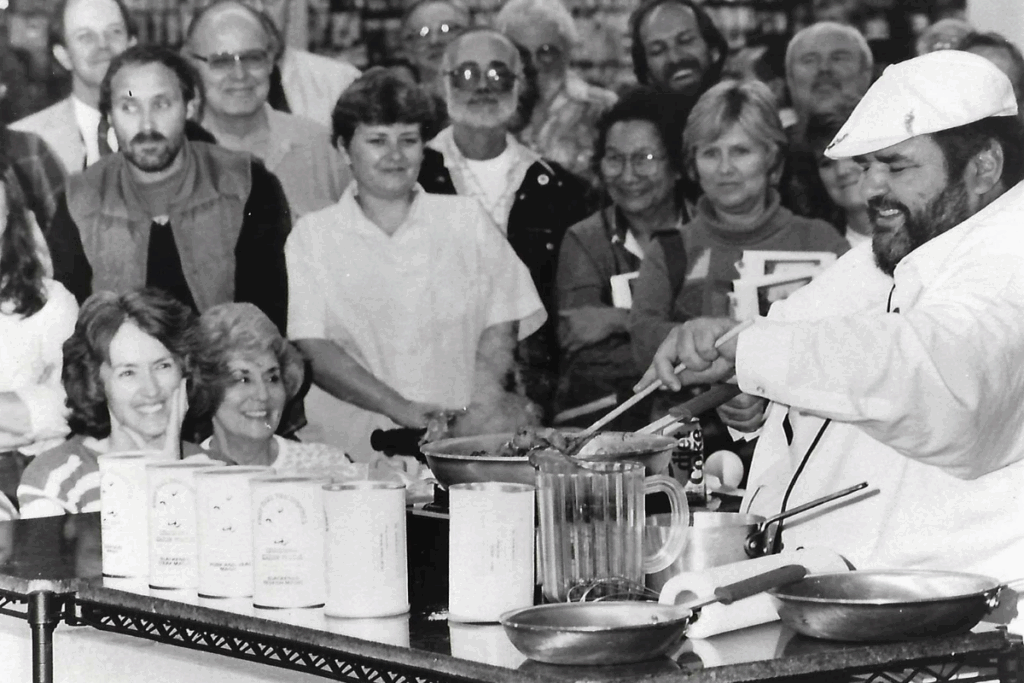 Chef Paul in a white uniform and hat, smiling, cooks at a demonstration. A diverse, happy crowd watches attentively. Various cooking items are visible.