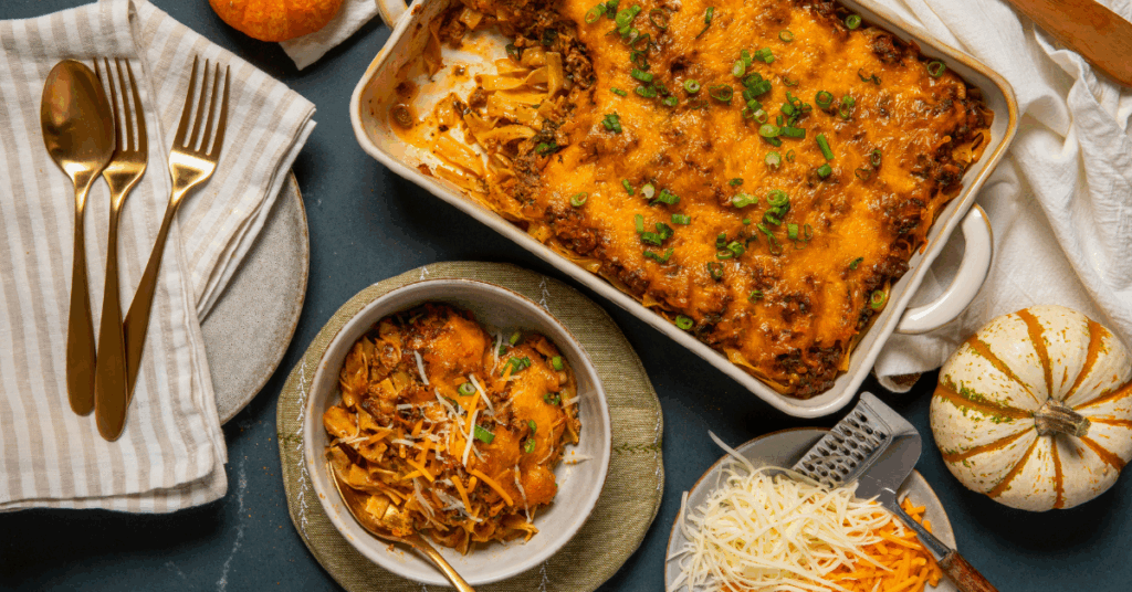 A baked cheese and pasta casserole with green onions in a dish. A served portion in a bowl with a fork; cheese and pumpkins nearby, evoking coziness.