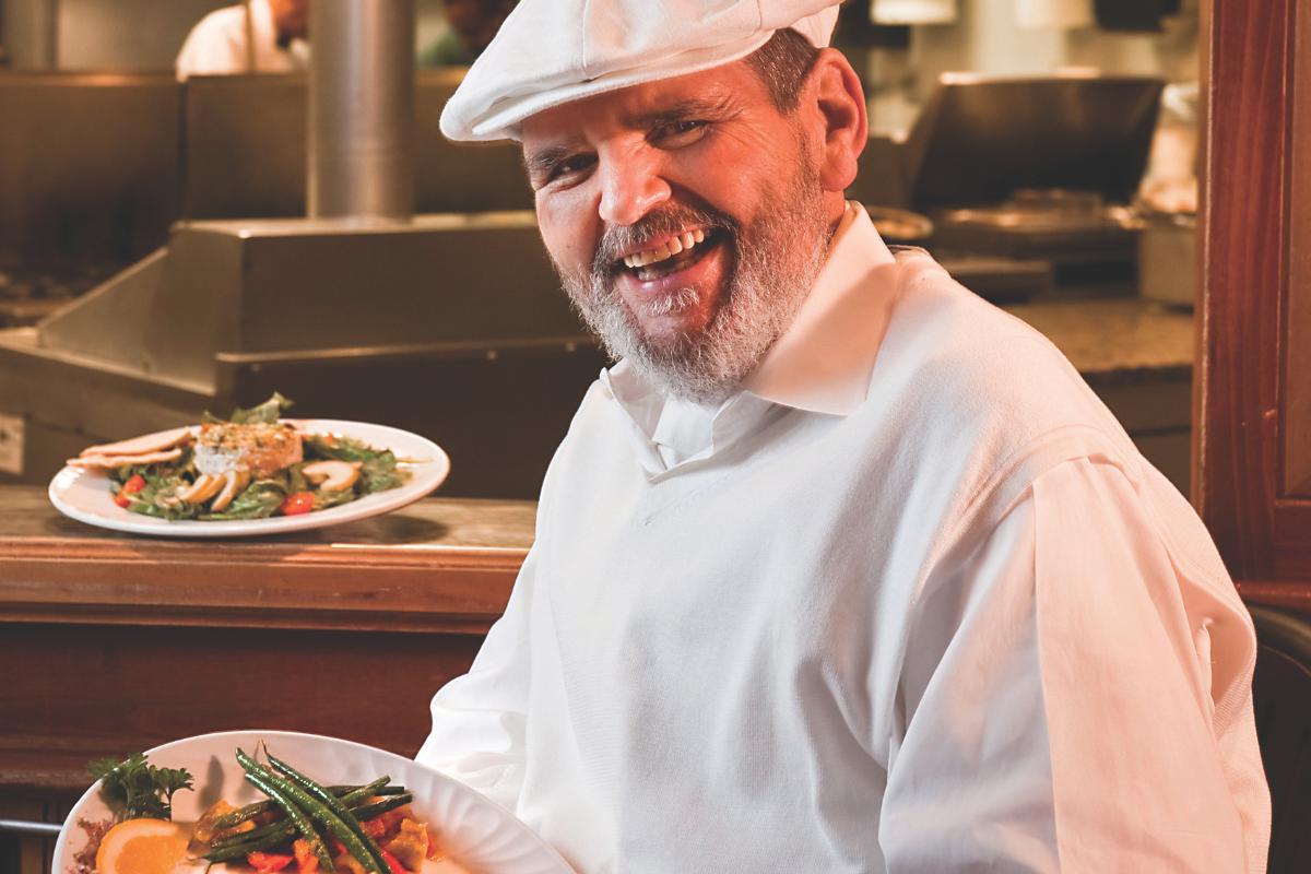 Chef Paul in a white outfit and cap smiles warmly while holding a plate of colorful vegetables and garnished dishes, exuding a joyful, welcoming atmosphere.