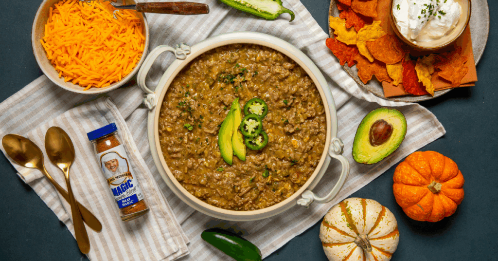 Comfort Food - A bowl of chili topped with jalapeño slices and avocado sits on a cloth, surrounded by cheddar cheese, chips with dip, spices, and small pumpkins.